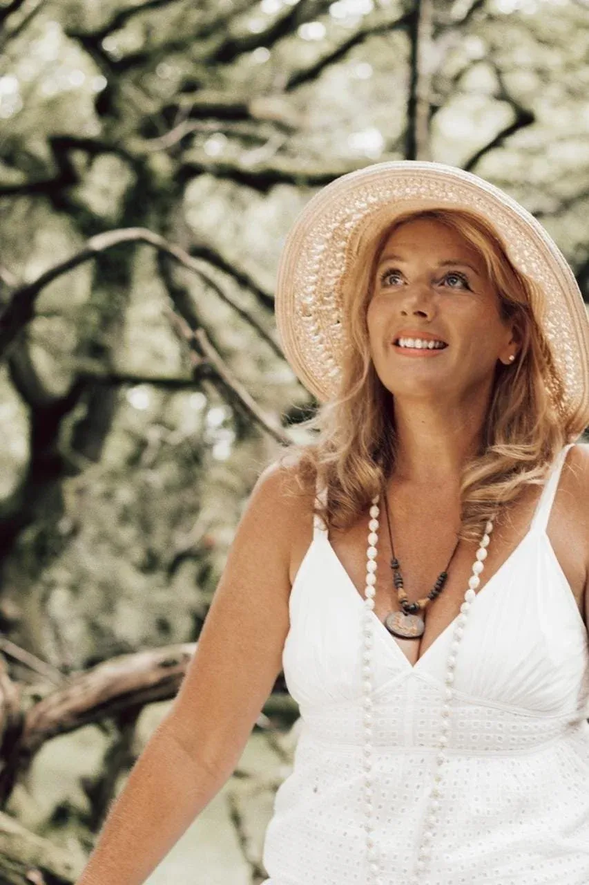 A woman with curly blonde hair wearing a wide-brimmed straw hat and a white sleeveless dress, standing outdoors among trees, smiling and looking up.