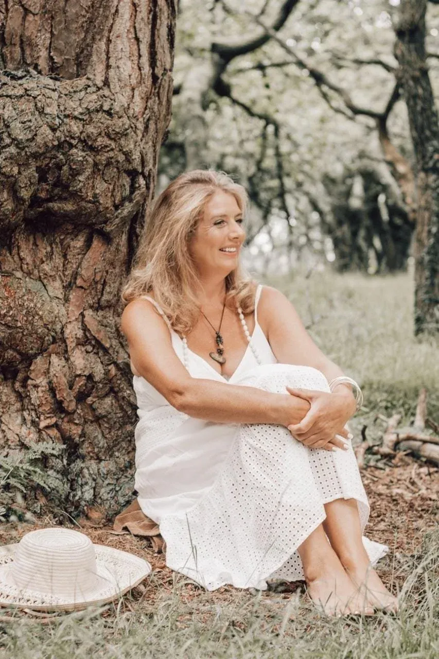 A woman with blonde hair wearing a white dress, sitting on the ground in a forest, leaning against a large tree, smiling, with a wide-brimmed hat on the ground nearby.