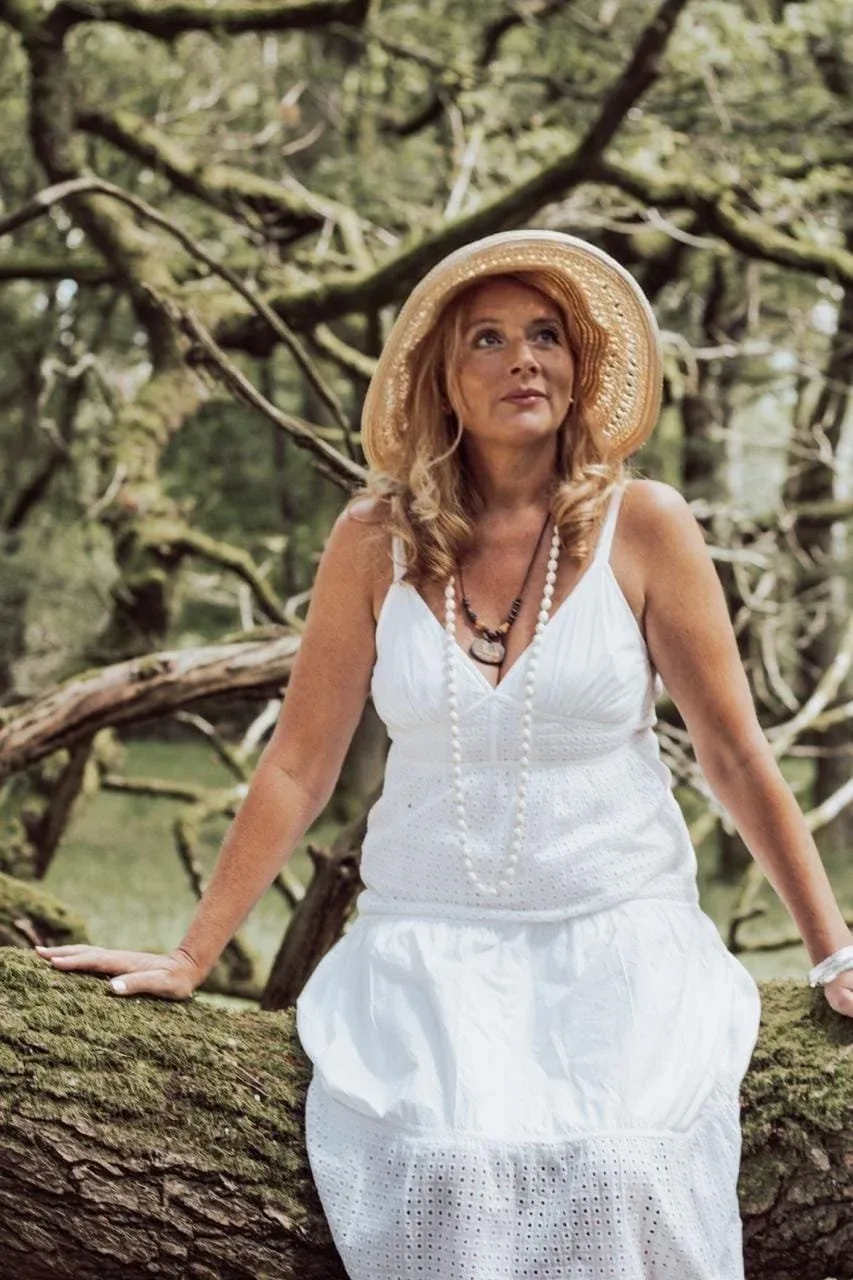 A woman in a white sundress and wide-brimmed straw hat sitting on a moss-covered log in a forest.